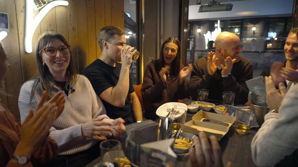 En grupp människor samlas vid ett bord på en bar eller restaurang, där några klappar händerna och en person håller ett glas. Det finns snacks och drycker på bordet, och stämningen verkar vara livlig och social.