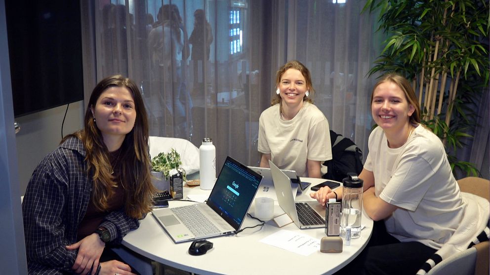 Tre kvinnor vid ett runt bord med datorer framför sig ler in i kameran