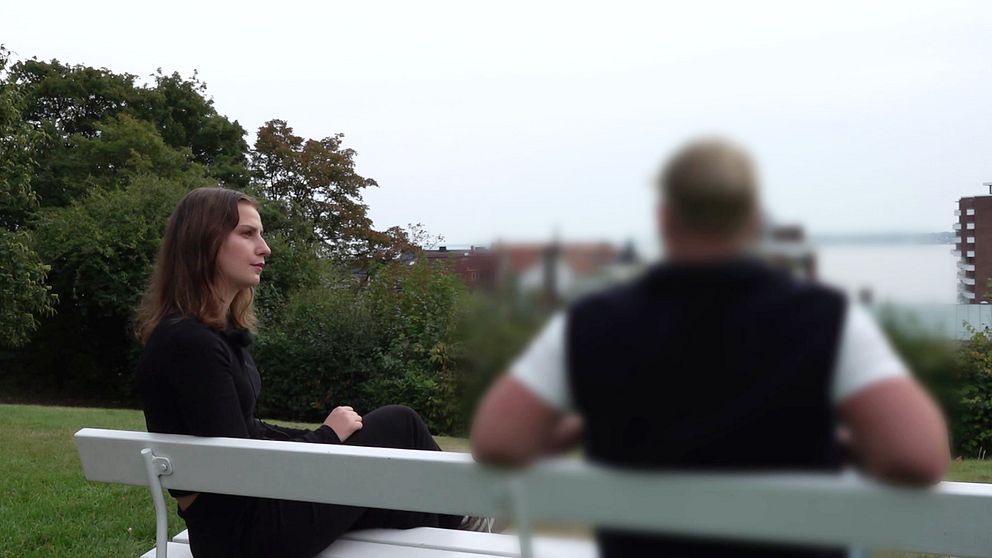 En kvinna sitter på en bänk i en park med grönska runt omkring. Hon är klädd i svart och ser ut över landskapet, medan en man med kort hår och svart/vit tröja sitter vänd bort från kameran. I bakgrunden syns en vy av staden och vatten.