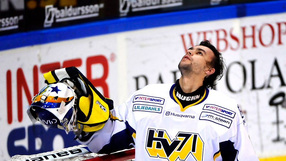 Målvakten Stefan Liv står vid målet med sina handskar och hjälm i ena handen, och ser uppåt med en känsla av lättnad eller triumf. I bakgrunden syns arenans läktare och reklamskyltar. Målramen är synlig i bildens vänstra del.