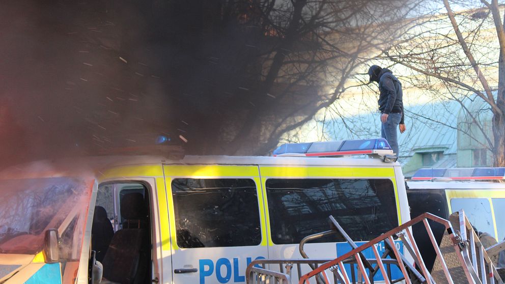 Man som står på polisbil, svart rök från brinnande fordon.