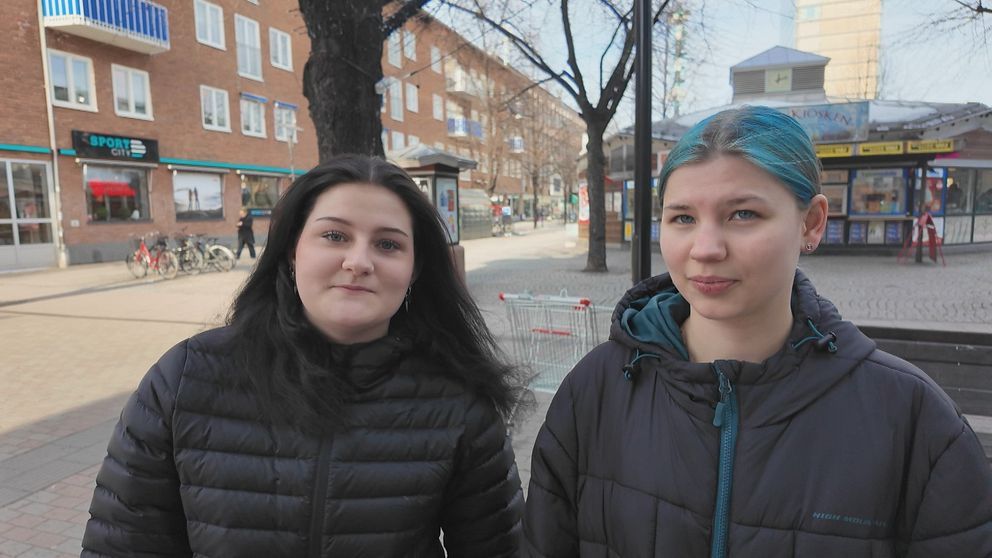 Elin Karlsson och Andrea Arklöf står på stadsgata i Skellefteå med butiker och ett träd i bakgrunden.