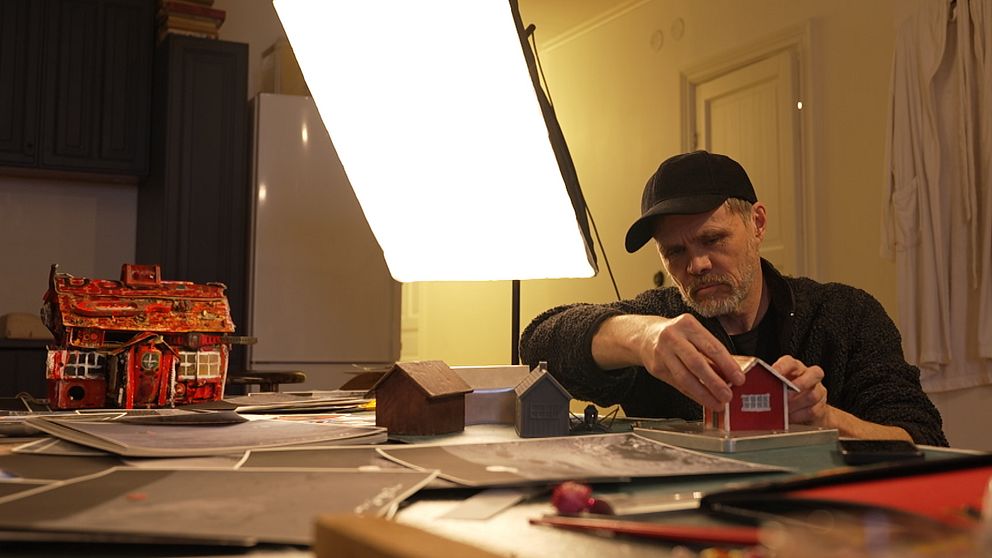 En person bygger modeller av hus vid ett bord med arbetslampor och material.