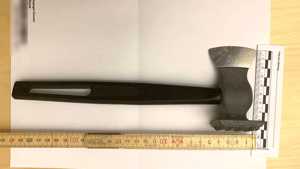 En yxa med svart handtag och silvrigt huvud ligger på ett bord. Bredvid yxan finns en linjal och ett måttband placerat för att ge en uppfattning om yxans storlek.