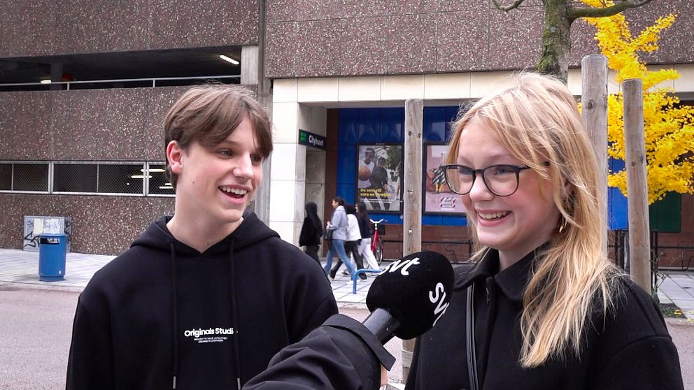En man och en kvinna står på en gata, en mikrofon syns, som tyder på en intervju. I bakgrunden syns en stadsbild med människor som promenerar och träd med höstlöv. Byggnaden bakom dem är av betong med stora fönster.