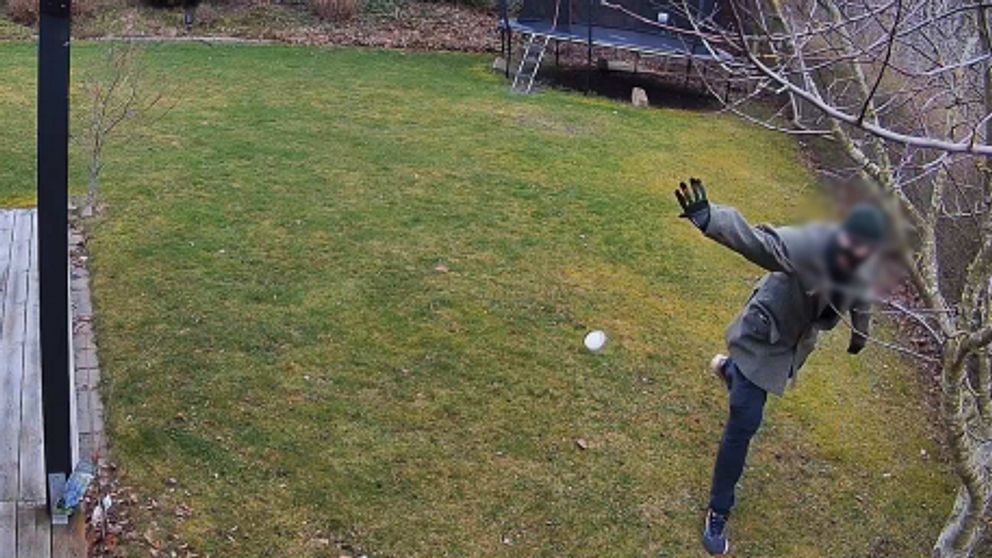 En person i en grå jacka och handskar kastar ett vitt föremål på en grön gräsmatta. I bakgrunden syns en trampolin och träd i ett kalt landskap.