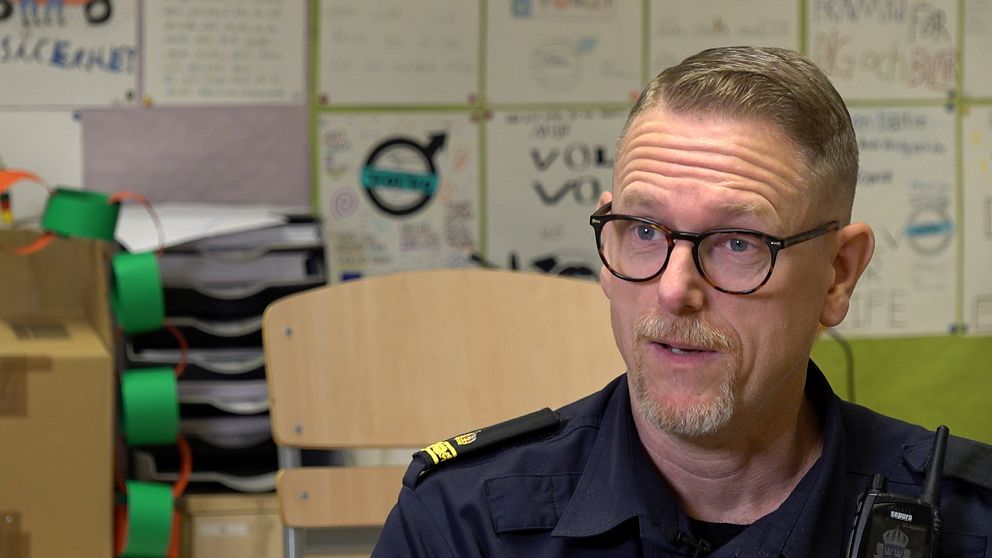 En person i uniform sitter i ett rum med synliga pappersplaner och konstverk på väggarna. Det finns också en stol och en stapel med papper i bakgrunden, vilket ger en känsla av en informell och kreativ miljö.