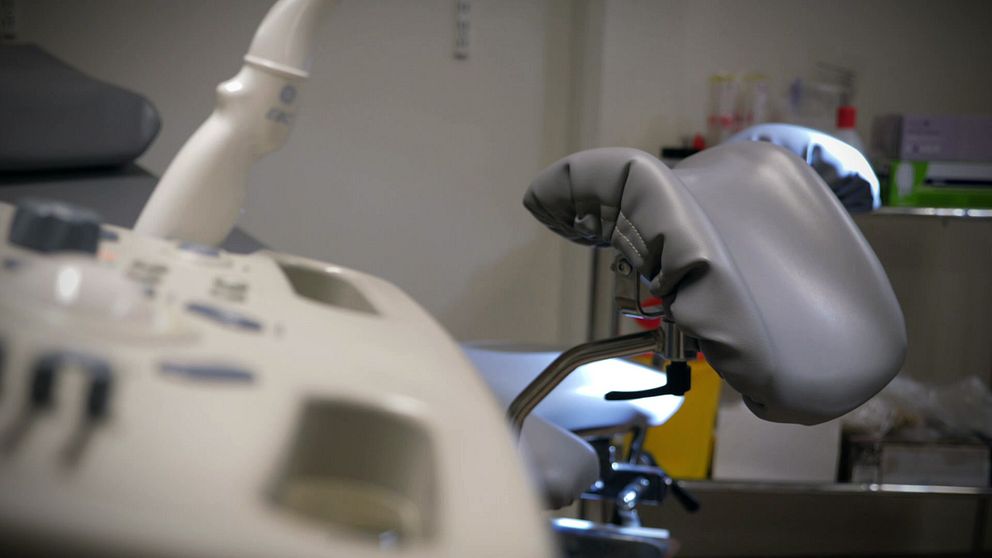 En bild som visar en del av en medicinsk utrustning, inklusive en vadderad stol med ett justerbart ryggstöd. I bakgrunden syns en arbetsyta med olika verktyg och utrustning i en klinisk miljö.