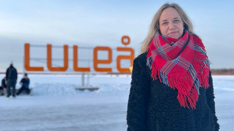 En kvinna står utomhus framför en stor skylt som visar texten ”Luleå”. Hon bär en svart jacka och en röd- och blåmönstrad halsduk. I bakgrunden syns vinterklädda människor och ett snöbelagt landskap.