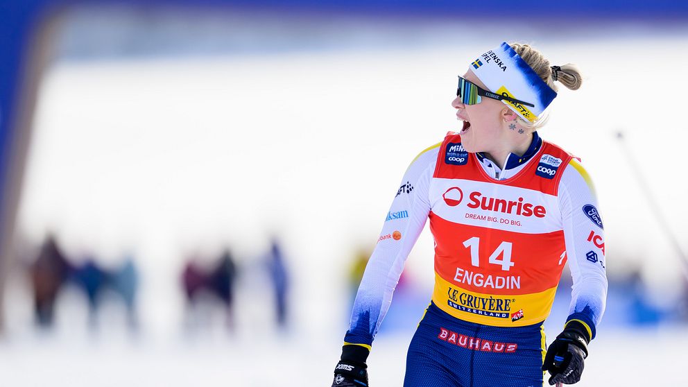 En kvinna deltar i en längdskidåkningstävling iförd nummer 14. Hon har en dräkt med sponsorloggor och håller i skidstavar, omgiven av ett snöklätt landskap. Bakgrunden är oskarp med flera människor synliga på avstånd.