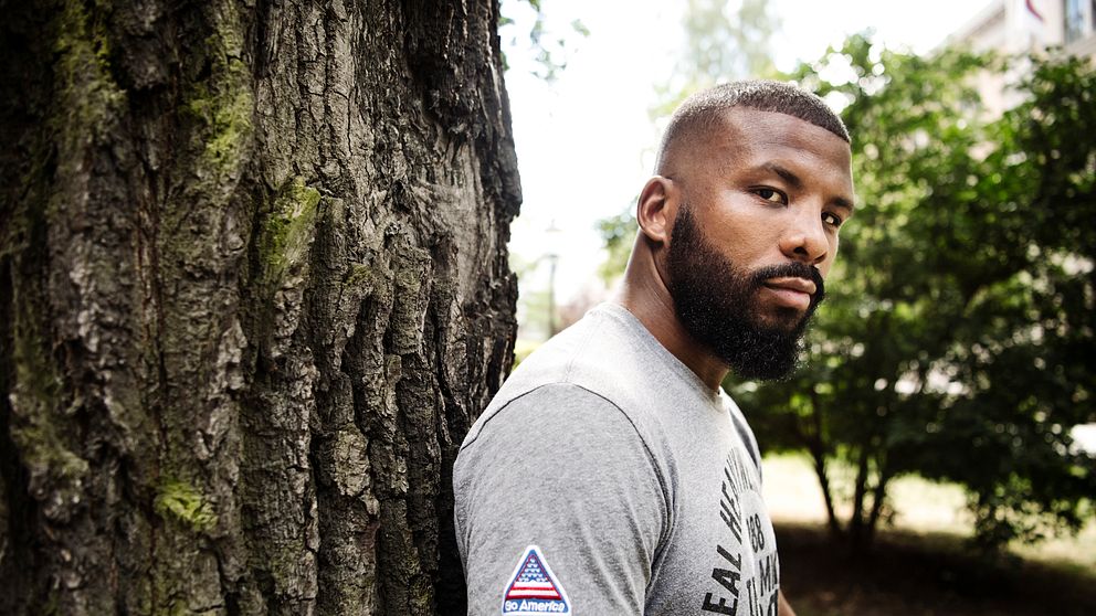 Man lutar sig mot ett träd i en parkmiljö, klädd i grå t-shirt med tryck.