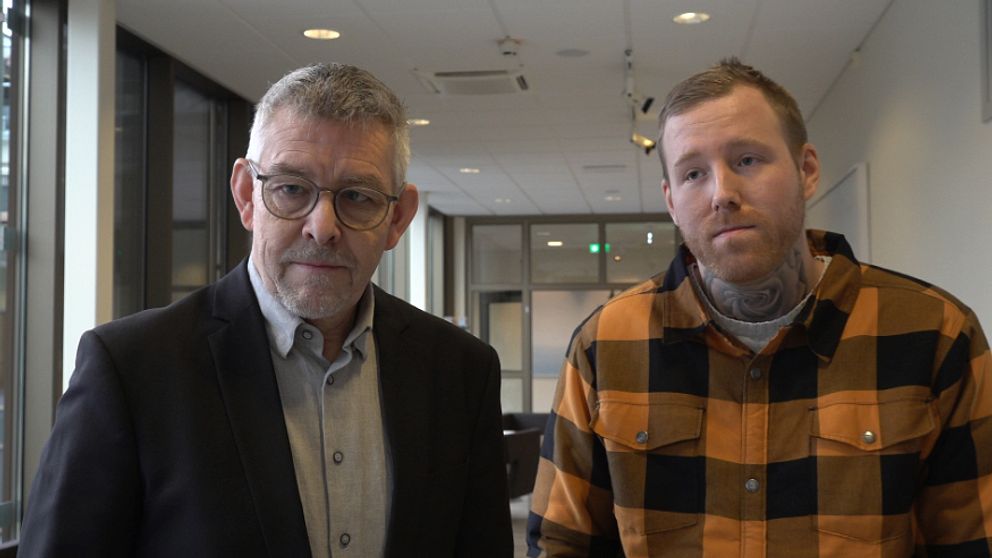 Två män står i en kontorsmiljö med moderna fönster i bakgrunden. Den första personen bär en mörk kavaj och en ljus skjorta, medan den andra personen har på sig en rutmönstrad skjorta i orange och svart. De ser mot kameran och verkar vara involverade i en diskussion eller presentation. De två männen är Arvikas kommundirektör Hans Karlsson och Robin Dahlén.