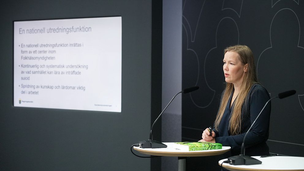 En kvinna står vid ett podium framför en skärm som visar en presentation.