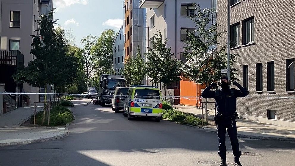 En polis står framför avspärrningar i ett bostadsområde med flerfamiljshus. Polisen håller för öronen.