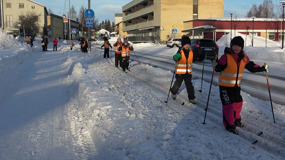 Barn på skidor i orangea västar åker skidor på led intill en bilväg.