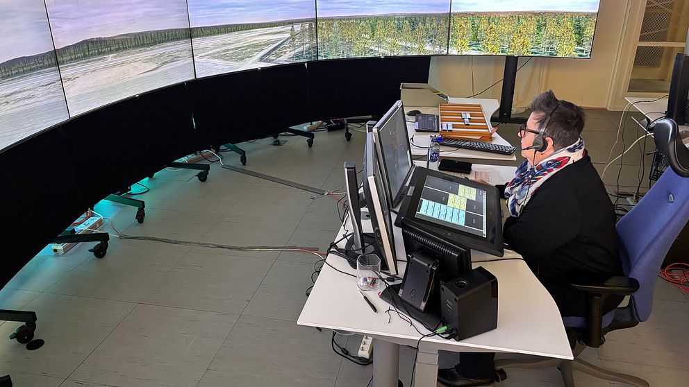 En person i arbetskläder sitter vid en datorstation med flera stora skärmar i en kontorsmiljö. Skärmarna visar en panoramautsikt över en flygplats som omges av skog och ett öppet landskap. Personen använder ett headset och ser koncentrerad ut medan de arbetar.