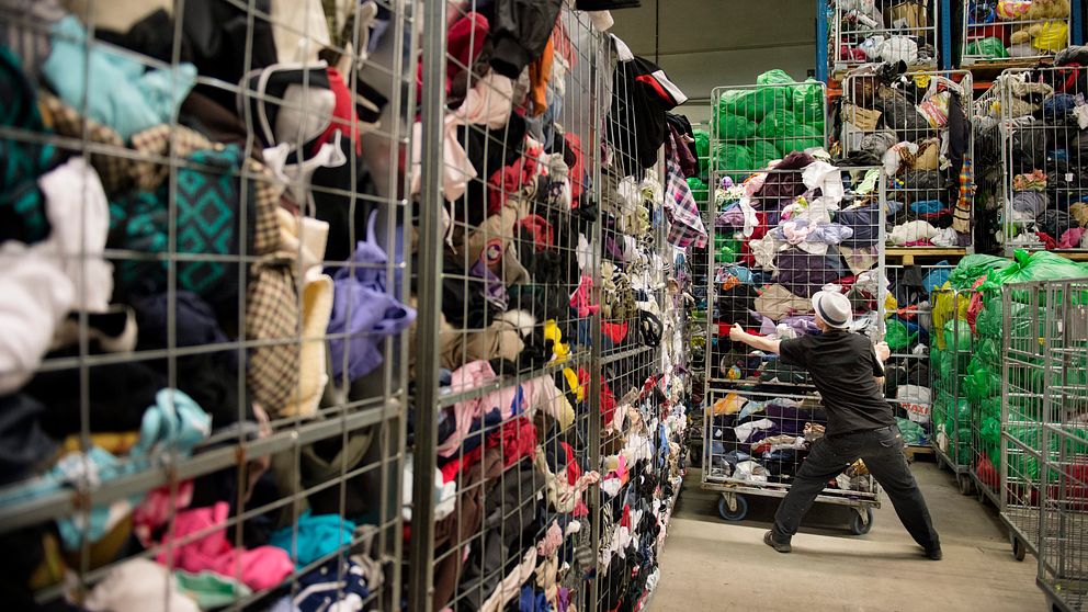 En person arbetar i en stor lagerlokal full med kläder som är sorterade i stora korgar. Korgarna står staplade på varandra och är fyllda med olika textilier i en mängd färger och mönster. Den anställde sträcker sig för att plocka upp eller sortera kläder från en av korgarna.