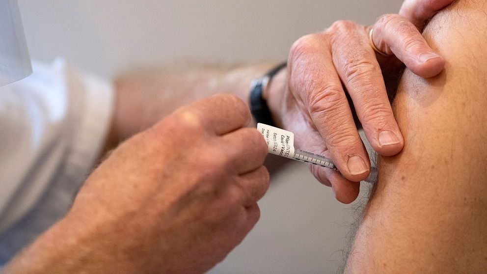 En bild på en spruta med vaccin mot covid 19 som sätts i överarmen på en man.