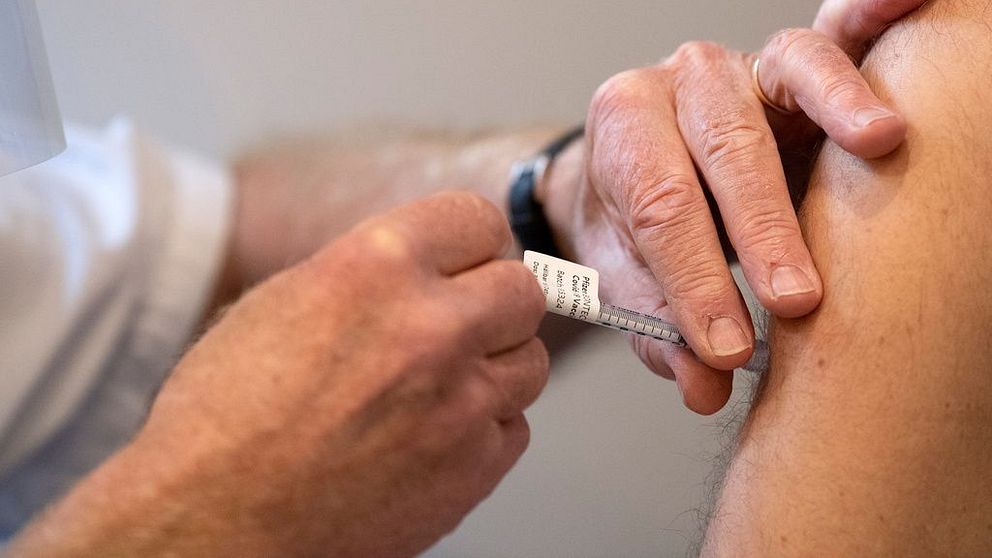 En bild på en spruta med vaccin mot covid 19 som sätts i överarmen på en man.