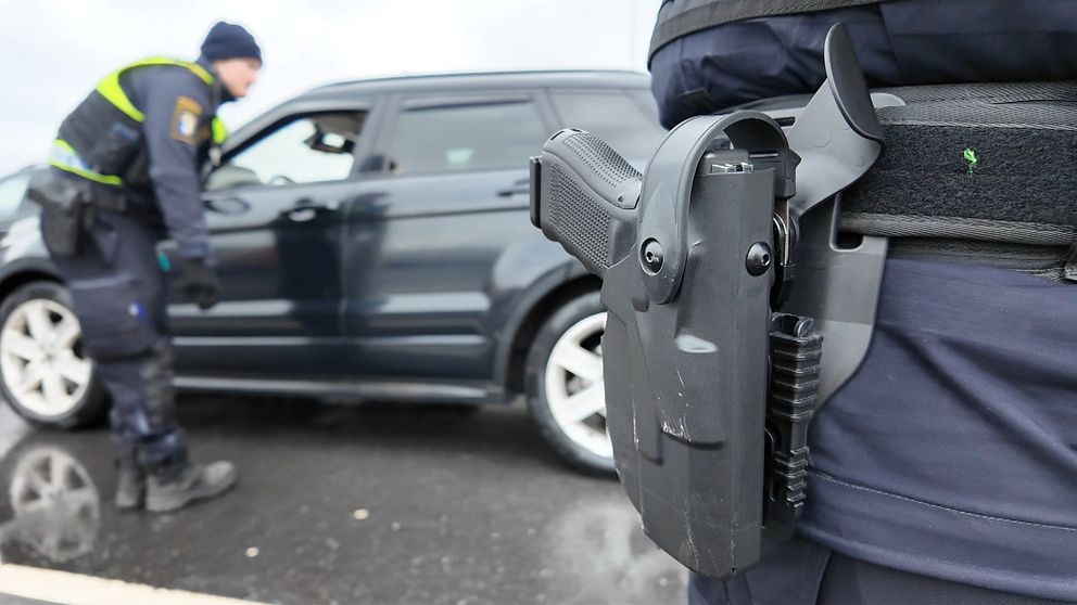En tulltjänsteman står vid en bil och pratar med föraren, medan en annan person är synlig i förgrunden med en pistol i hölster. Scenen visar en ordningskontroll på en väg.