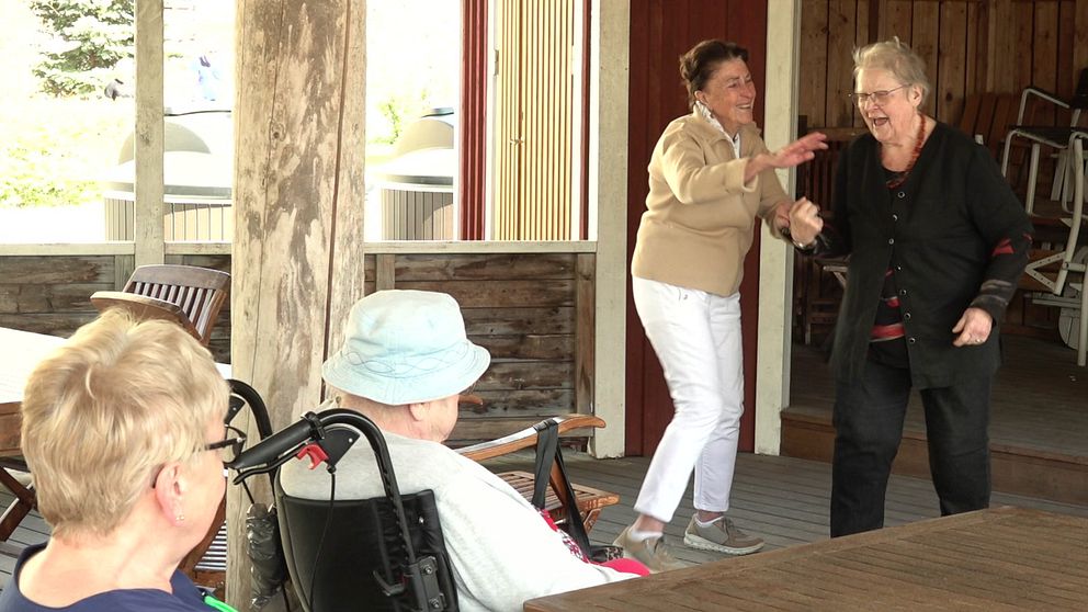 Två äldre kvinnor dansar på något som kan vara en dansbana, I förgrunden ser man en person i rullstol med ett hatt på huvudet och ytterligare ett huvud bakifrån (kvinna, kortklippt blont hår, glasögon).