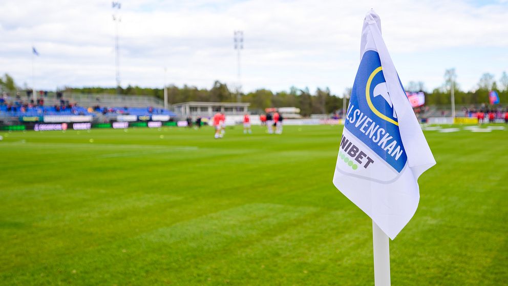 Hörnflagga med ”Allsvenskan” på fotbollsplan, spelare värmer upp i bakgrunden.