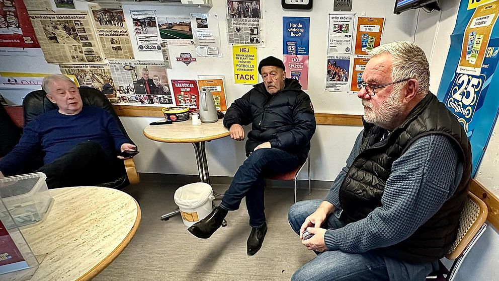 Tre äldre män sitter och pratar i ett rum med lottoaffischer på väggarna.