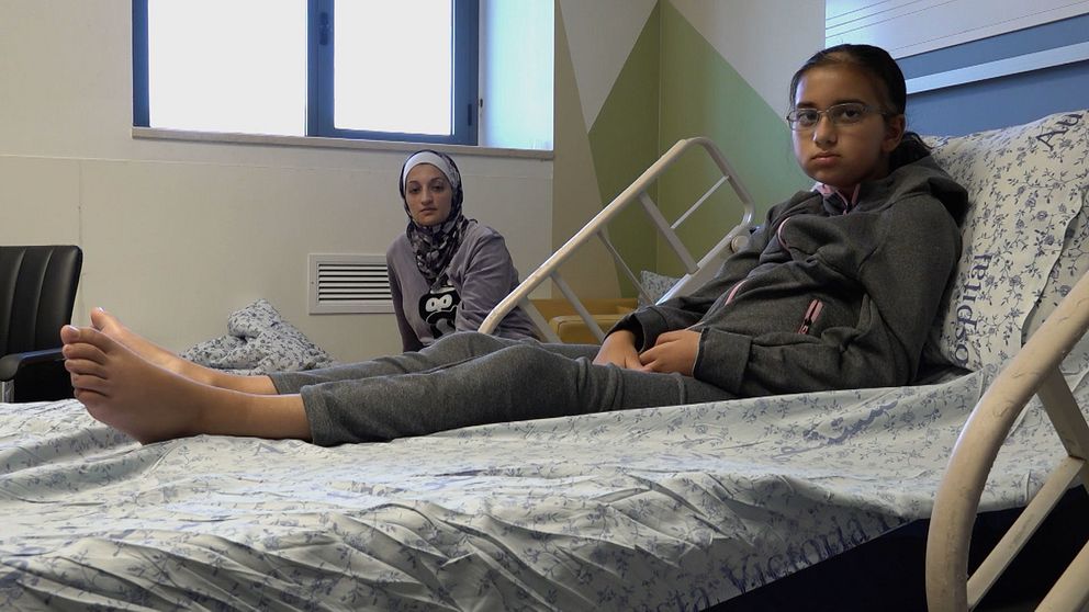 Amira ligger i sjukhussäng, hennes mamma sitter bredvid sängen.