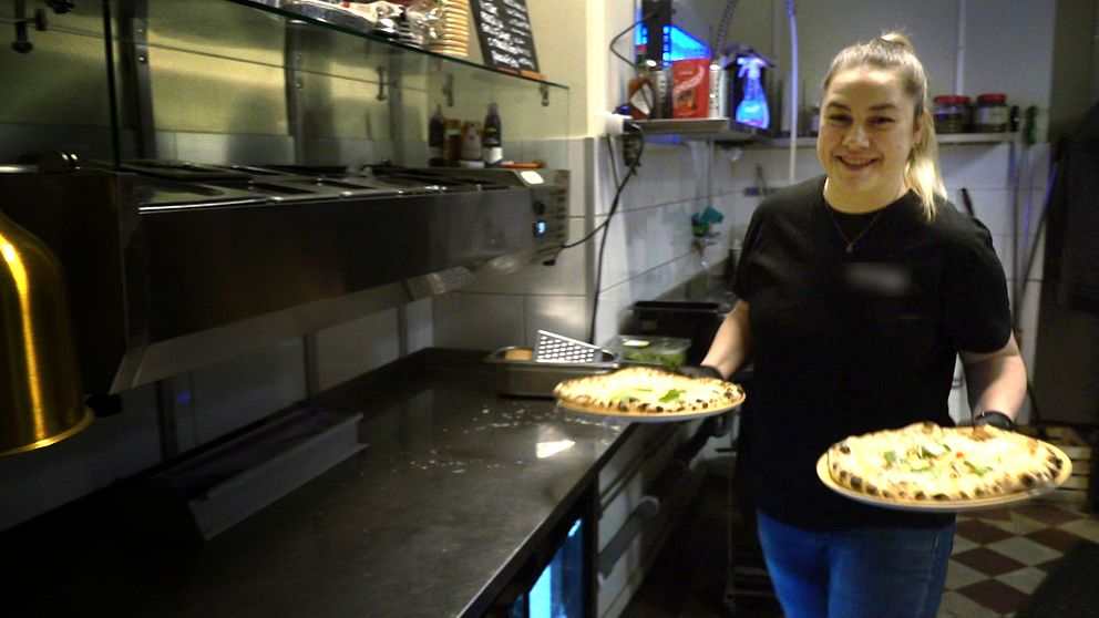 Kvinna bär två pizzor i restaurangkök.