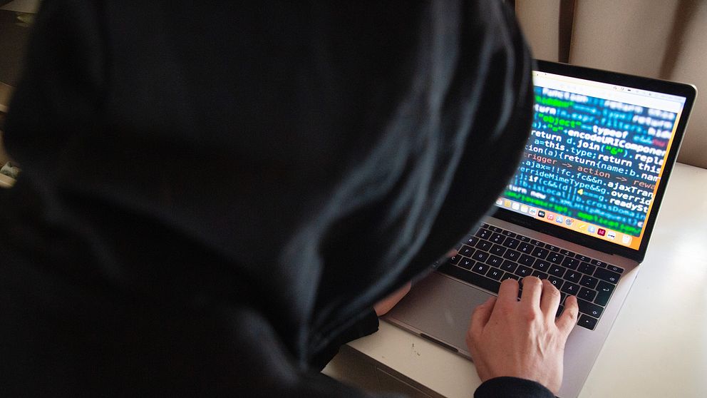 En person i luvtröja skriver på en dator som har massor av koder på skärmen.