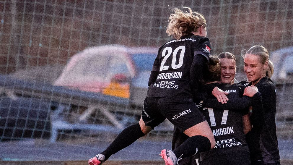 En grupp kvinnliga fotbollsspelare i svarta tröjor firar ett mål genom att omfamna varandra, medan en spelare i vit tröja observerar från sidan. Bakgrunden visar en fotbollsplan med gräs och en samtida infrastruktur.