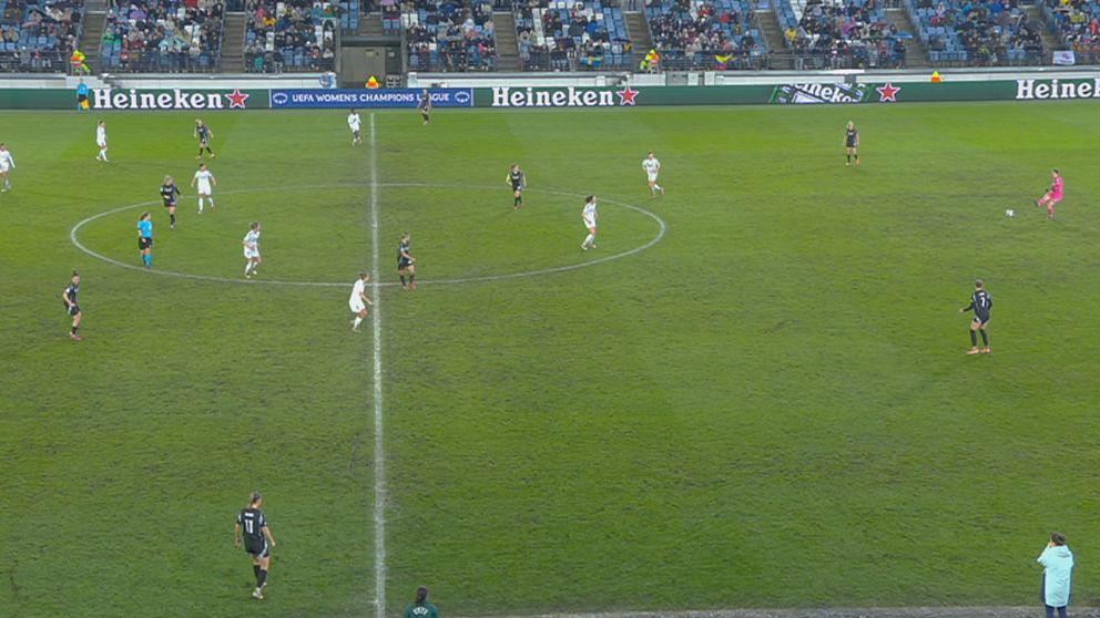 Fotbollsmatch på plan med spelare från två lag, åskådare i bakgrunden.