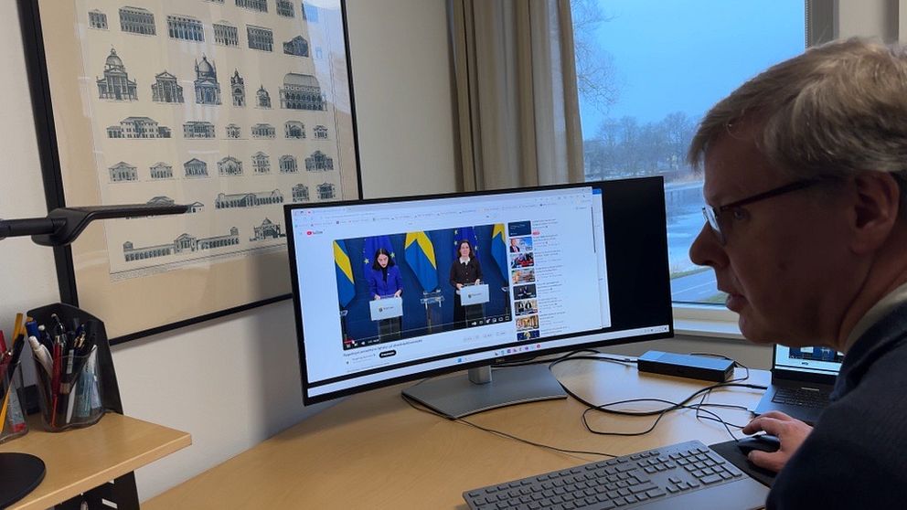 En person sitter vid en dator och tittar på en livestreamad presskonferens med svenska flaggor i bakgrunden. Skärmen visar två talare som står vid podium med mikrofoner under presentationen. I rummet syns även en teckning av byggnader på väggen och skrivmaterial på skrivbordet.