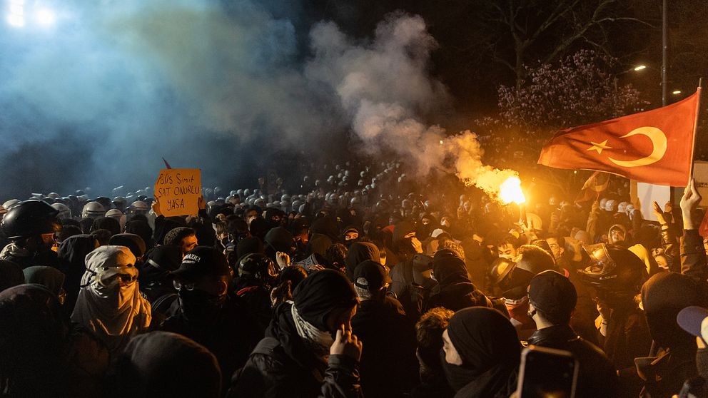 Folkmassa protesterar med turkisk flagga och facklor, rök fyller natthimlen.