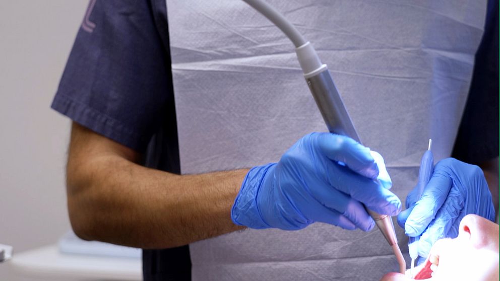 En tandläkare i blå skyddshandskar utför en behandling på en patient, med ett dentalverktyg riktat mot patientens mun. Bakgrunden visar en sjukvårdsinrättning med en tandläkarstol synlig.