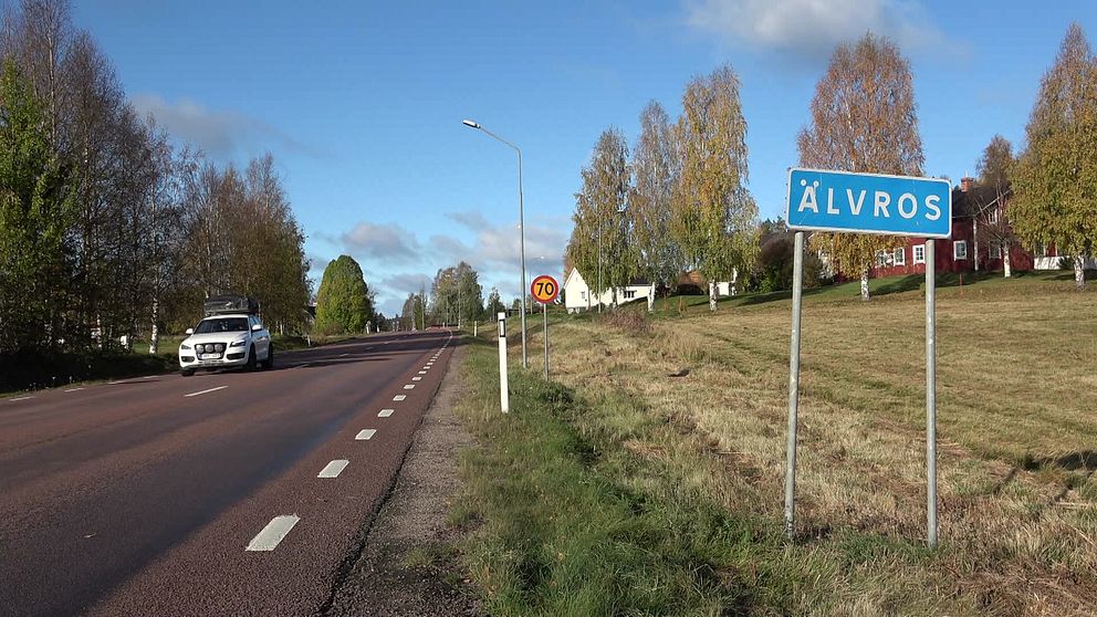 Vägskylt som visar namnet ”Älvros” till vänster, med en förbipasserande bil och omgivande träd och hus. Motivet visar en landsväg omgiven av grönska och hus i bakgrunden. En hastighetsbegränsningsskylt med 70 km/h syns också.