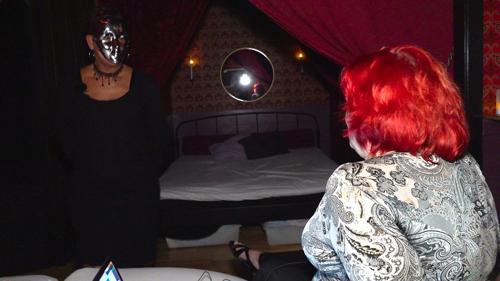 Arrangör till swingersklubb sitter på säng tillsammans med annan besökare med mask på.