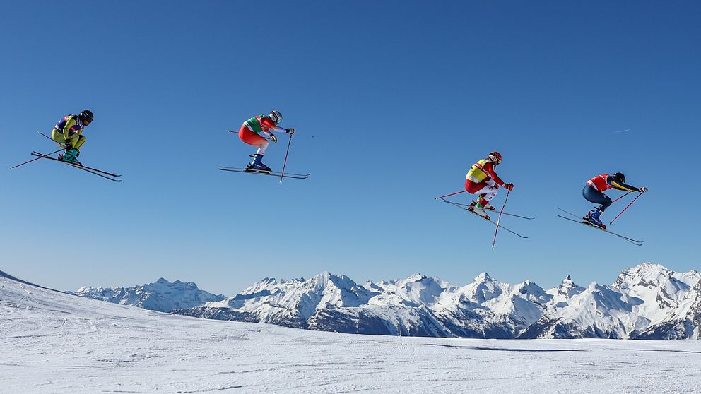 Fyra skidåkare hoppar i luften samtidigt under en skicross-tävling, med snötäckta berg i bakgrunden och klarblå himmel ovanför.