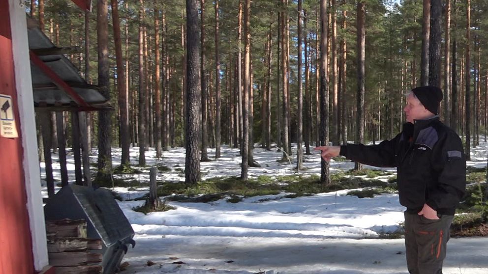 Man som står och pekar mot en vedbod där det står en skopa från en grävmaskin.