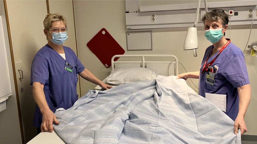 Två undersköterskor i lila ställ bäddar sjukhussäng. De tittar mot kameran och bär munskydd.