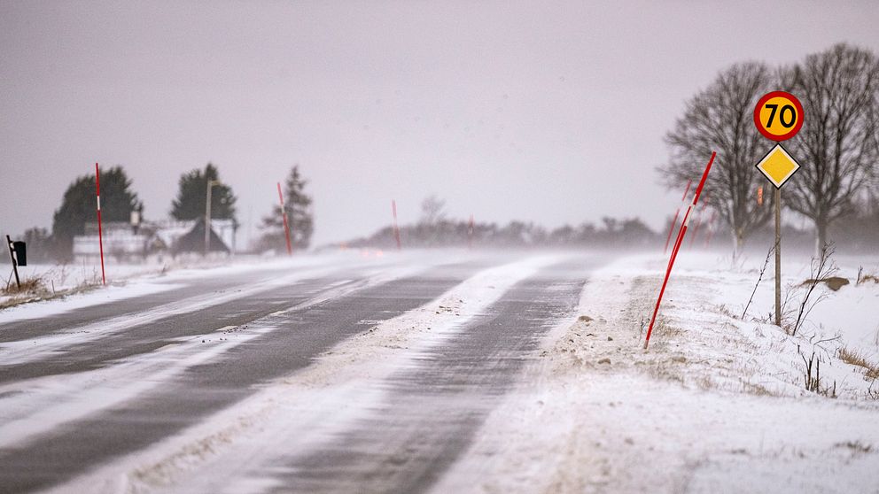 Snöig landsväg med vind och hastighetsskylt på 70 km/h, buskar och träd i bakgrunden.
