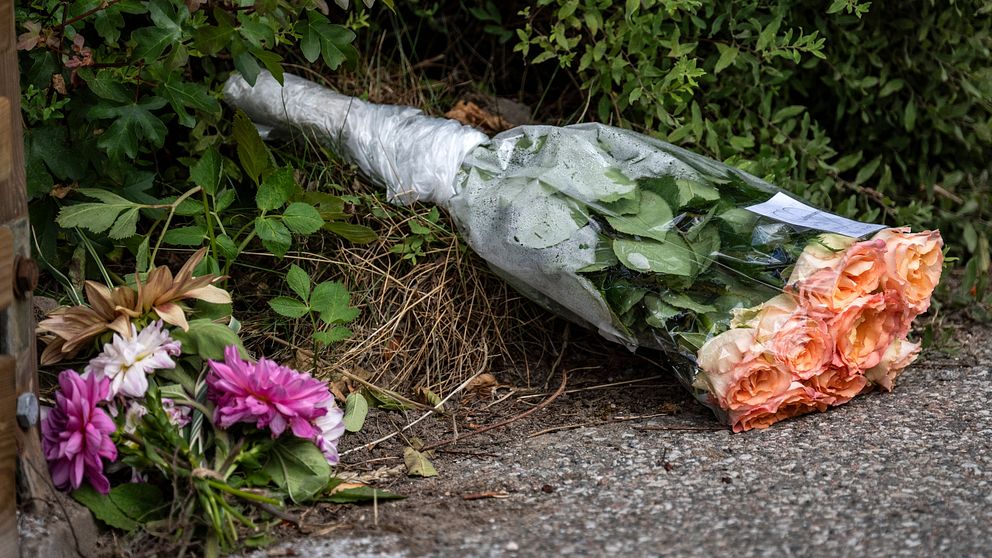 En bukett med rosor i plastfolie ligger på marken bredvid en annan blomsterrangering av olika färger, omgiven av grönska.