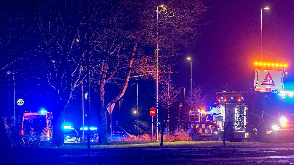 Utryckningsfordon med blinkande blåljus är uppställda längs en väg på natten. Ett fordon har även en skylt med varningsblinkers för att dirigera trafiken.
