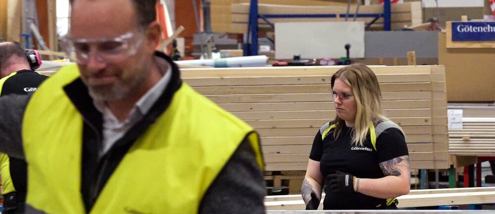En bild från fabriksmiljö. Till vänster en oskarp man i gul väst och skyddsglasögon, till höger en ljushårig tjej som står och arbetar.