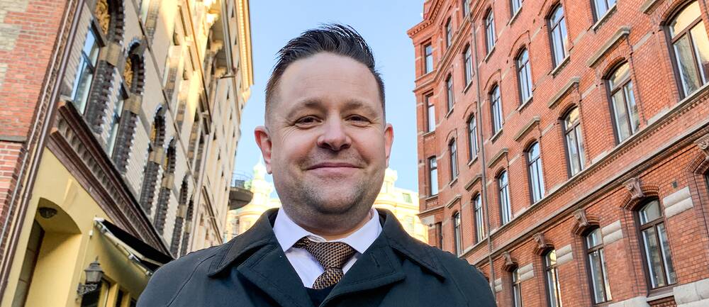 En bild på en Johannes Hulter som ser glad ut, och står framför två gamla tegelbyggnader i Göteborg.