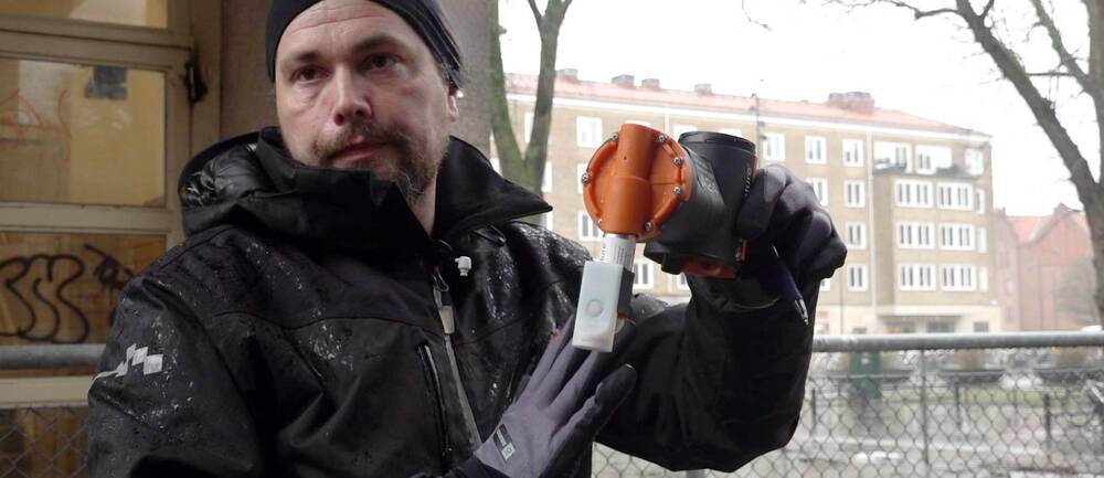 Skadedjurstekniker Anders Israelsson visar upp en råttfälla. Han står utomhus framför en byggnad och ett staket.
