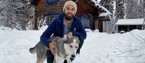 Youtubern Kalle Flodin sitter på huk i snön och håller sin huskey Tuss i famnen. I bakgrunden ser vi hans timmerstuga.