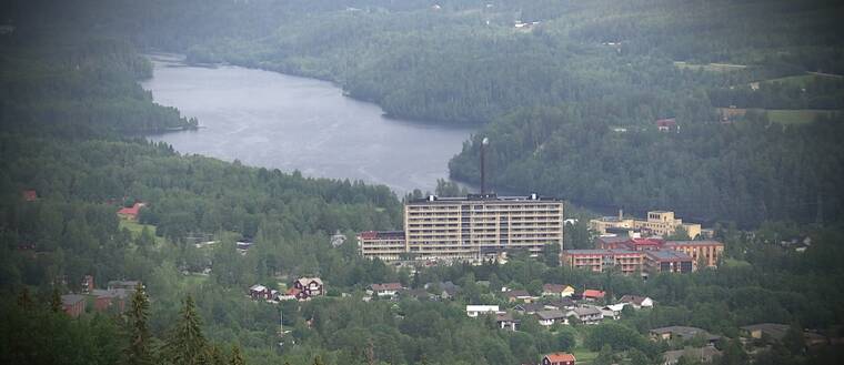 vy över bebyggelse omgiven av skog, sjö bakom. Äldre trähus och stora komplex