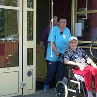 Olena Hrytsanchuk med en kollega och brukare – en gammal kvinna i rullstol – på väg ut på promenad. Olena är klädd i blått och har mörkt lockigt hår och hennes kollega har på sig lila kläder och långt ljust hår.