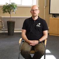 En man i bruna chinos, svart piké sitter på en stol i ett konferensrum. Han heter martin eneborg